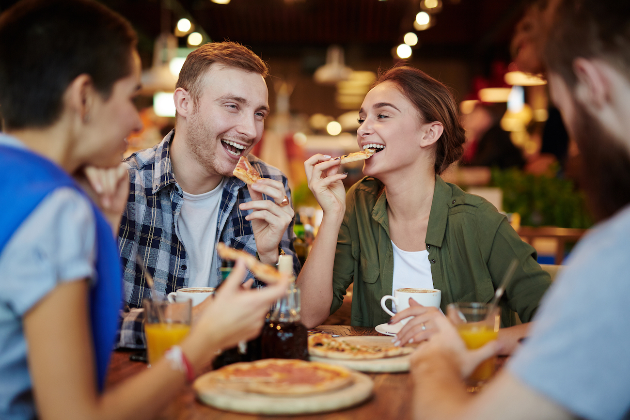 Group of cheerful friends eating delicious pizza and chatting animatedly while gathered in lovely small cafe, waist-up portrait