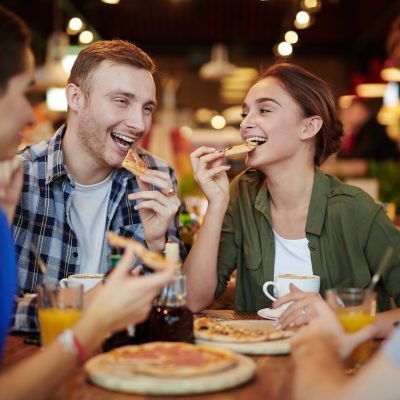 Group of cheerful friends eating delicious pizza and chatting animatedly while gathered in lovely small cafe, waist-up portrait